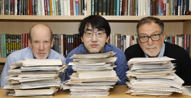 Three men rest their chins of stacks of documents piled on a desk