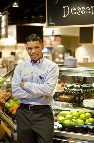 A man, smiling slightly and wearing a blue dress shirt and slacks, leans against a deli counter display filled with fresh fruit and pastry