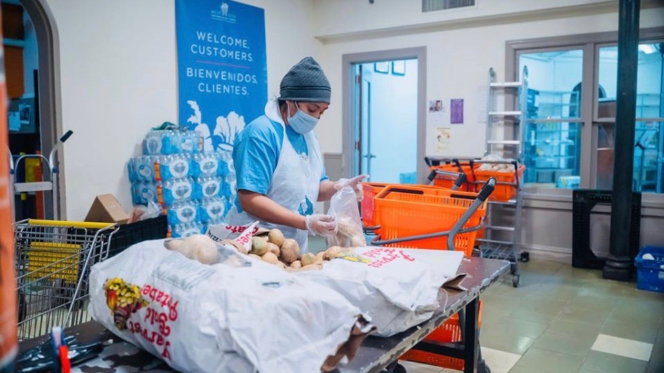 A worker in a hat, mask, gloves, and apron sorts through giant bags of potatoes at a food pantry.