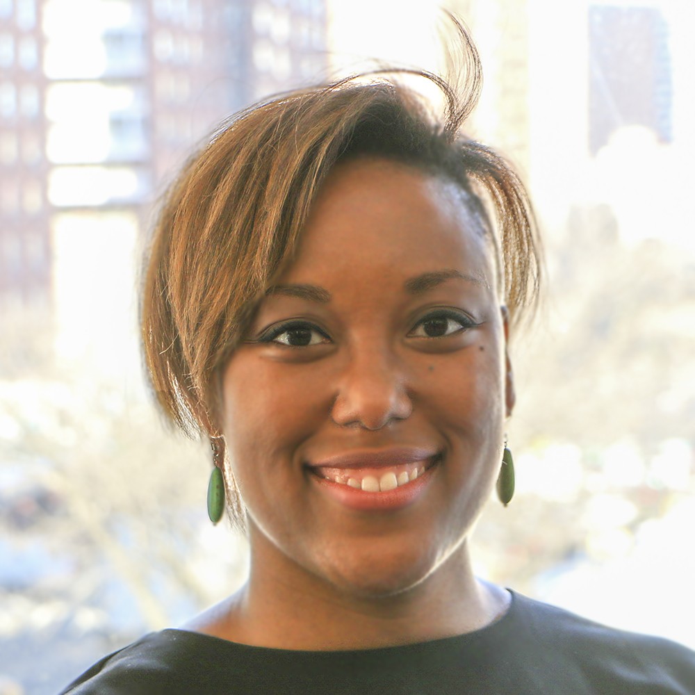 A woman, wearing a black boat-neck blouse and jade green earrings, miles into the camera