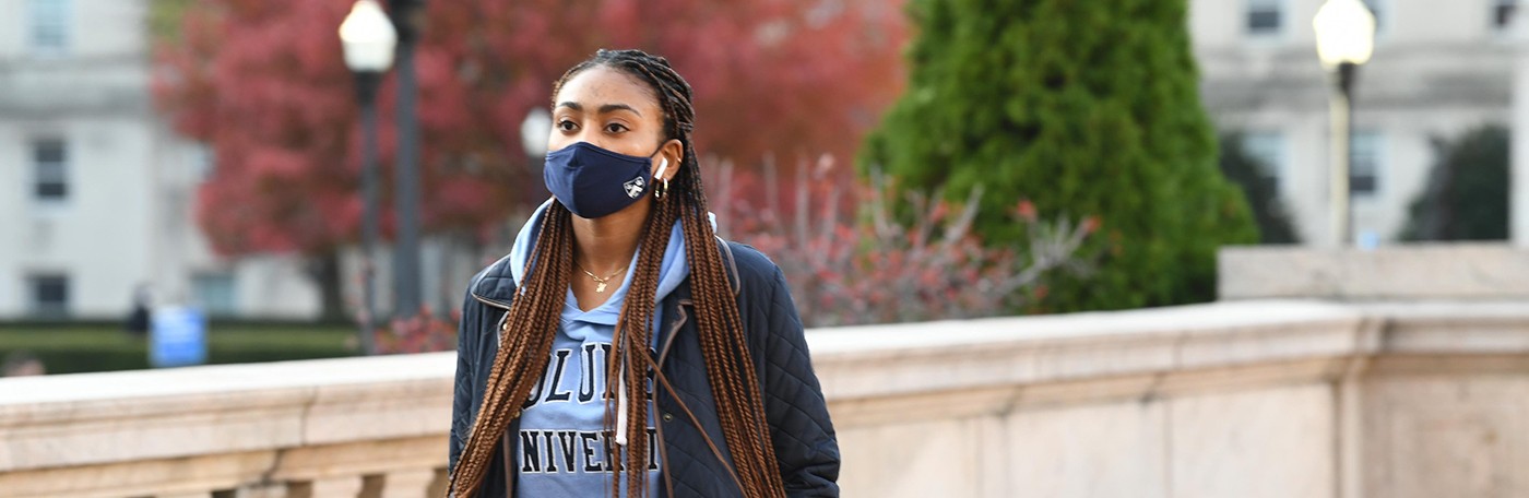 A Columbia student in a blue sweatshirt and face mask walks across Morningside campus.