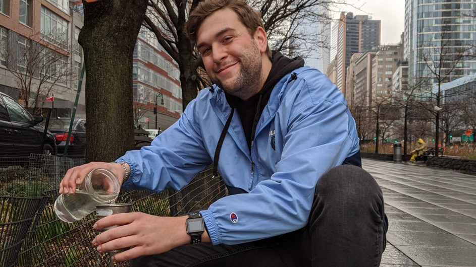 A young blonde man with a light beard and wearing a Columbia Athletics jacket, crouches near a fenced-in tree while pouring liquid from a mason jar into a tin container for testing.