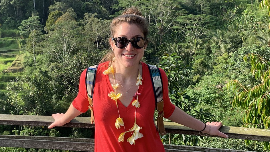 A young white woman with light brown hair in a bun is wearing sunglasses, a red v-neck t-shirt, and a yellow floral lei while standing against the rail of a wooden bridge overlooking lush green treetops.