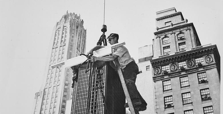 A man stands stop a ladder, untying a rope that is securing a model of the Seagram Building from a pulley.