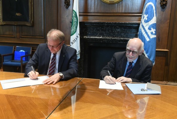 Two men signing papers on a wooden table in front of flags