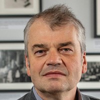 Dmitri Basov: A man with closely cropped grey hair staring into the camera with framed black and white photos behind him.