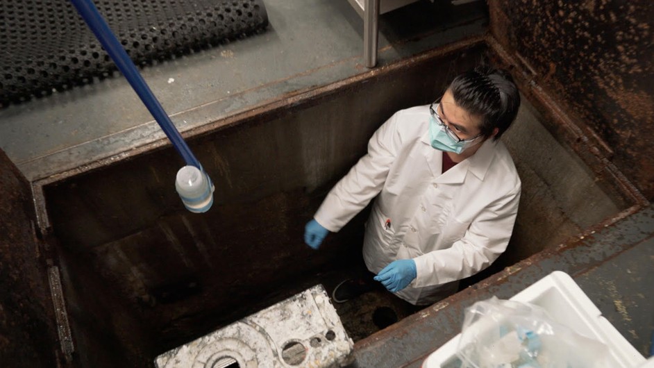 Person with brown hair, glasses, blue mask and gloves, and white jacket stands in hole in dark room