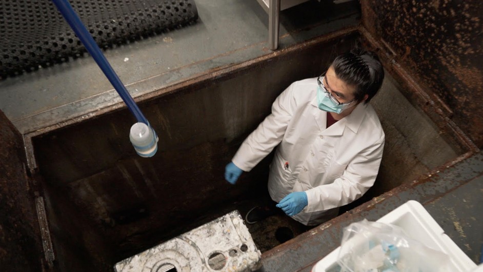 Chengua Long, a postdoc researcher in Kartik Chandran's lab, takes wastewater samples at a Columbia dormitory to be tested for the presence of SAR-CoV-2. Photo: Columbia Engineering Person with brown hair, glasses, blue mask and gloves, and white jacket stands in hole in dark room