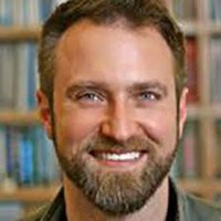 Headshot of Caucasian man with short brown hair and beard smiling with bookcases behind him