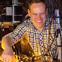 Sebastian Will headshot of white man smiling with checked shirt with background of yellow-lit lab equipment
