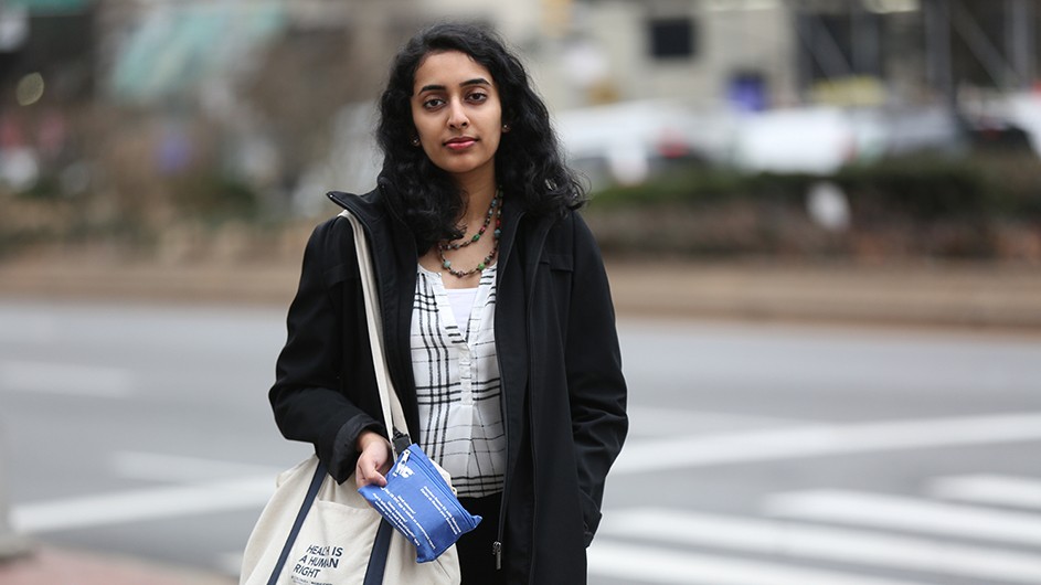 Young woman with black hair wearing a black jacket in a blurred urban setting. She carries a blue kit strapped to a while shoulder bag.