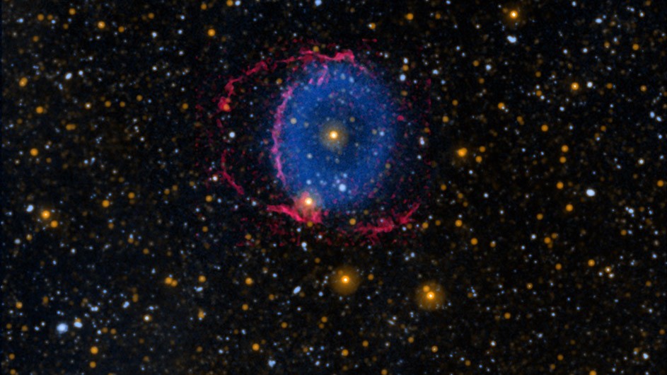 The Blue Ring Nebula is thought to be a short-lived phase during the merger of two stars. When the stars collided, debris was flung outward to form two cones that fanned outward from the stars in opposite directions. The area where the bases of the cones overlap, color-coded blue, represent ultraviolet light. The edges of the two cones are color-coded pink, representing visible light. Image credit: NASA/JPL-Caltech/M. Siebert (Carnegie Institution for Science)/K. Hoadley/Caltech/GALEX team Black sky with hundreds of small stars and at center a blue donut-shaped ring with red lines surrounding it