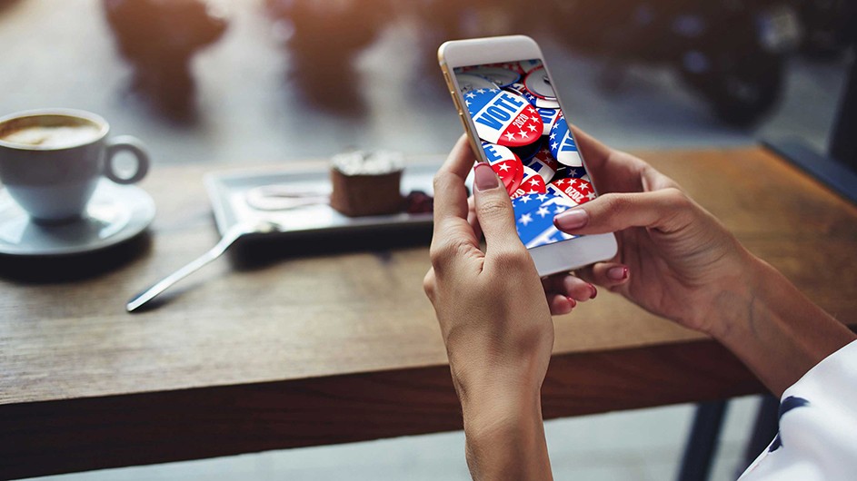 Hands hold a cellphone with image of red, white and blue VOTE buttons; a piece of cake with plate and spoon, plus white cup of expresso lies on a brown table