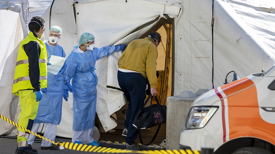 Yellow-masked EMT behind two blue-uniformed clinicians entering white tent