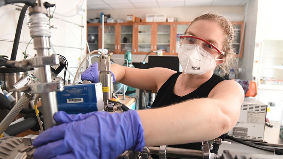 woman with pulled back hair red goggles, purple gloves and white mask working on equipment