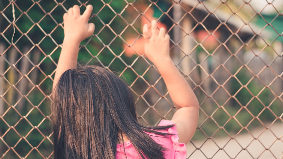girl in pink dress with long brown hair and back to us holds arms raised on steel fence with criss-crosses