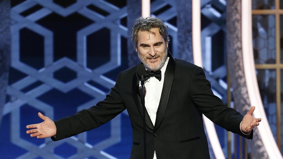 The actor Joaquin Phoenix, at this year's Golden Globe Awards, has vowed to recycle his tuxedo throughout the awards season. Middle-aged man with grey hair and beard smiling wearing black and white tux. Hands opened wide. Against light blue background