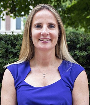 upper body woman with blond hair and purple sleeveless dress with green bushes on background