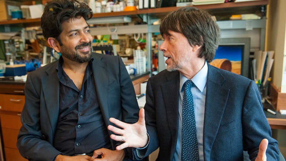 Darker skinned man on left with black hair and beard, smiling, has turned sideways to speak with shorter white man with brown hair and grey beard who is wearing a tie. Both men in sports jackets.