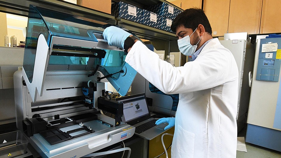 Man with dark hair, beard, mask, gloves and white lab coat is turned sideways lifting the cover of a large white and plexi glass blue machine