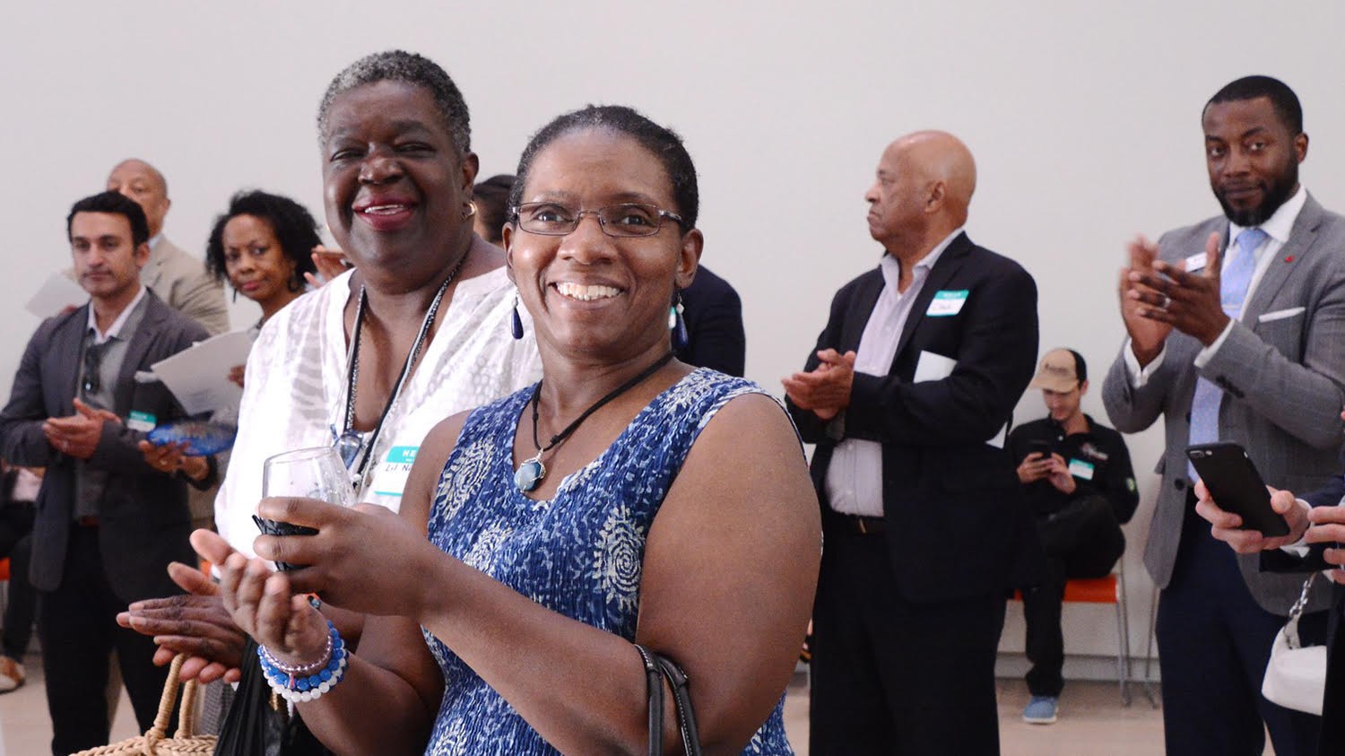 Lil Nickelson (left), a Columbia Community Scholar and a writer for Harlem World Magazine, and Ramona Prioleau (right) of FraMiaTi, a philanthropic natural skincare company based in Harlem, are all smiles at the anniversary reception. Both women are SBDC alumnae. Two women smiling and clapping in the center among a crowd of people.