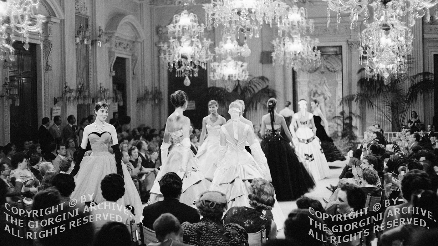 A 1950s Giovanni Battista Giorgini fashion show at the Sala Bianca in the Palazzo Pitti in Florence, Italy. Woman in ballgowns are walking a runway at a fashion show surrounded by a large audience, all of this taking place in an ornate room with glass chandeliers.