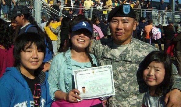 An Asian family of four with a woman in the center holding up a certificate next to a uniformed man