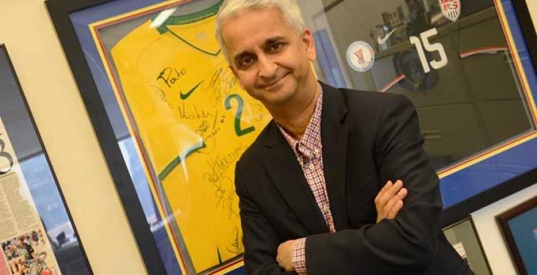 Sunil Gulati stands with arms folded smiling into camera wearing a dark blazer with red black and white plaid button down shirt