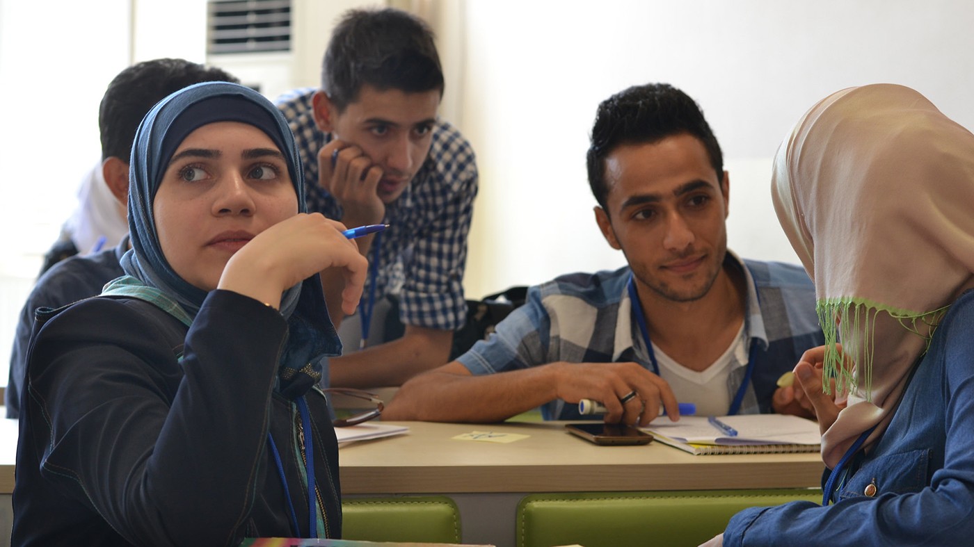 Photo of five refugee students, including two women in scarves, gathered around a desk