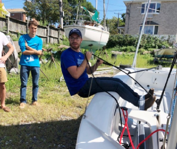 Treatman showing a sailing maneuver to the Columbia sailing team. David Treatman: A man wearing jeans, baseball cap and a T-shirt on a sailboat holding a rope and leaning backward.