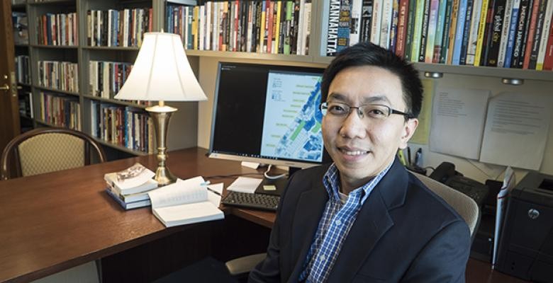Van Tran wearing eyeglasses wth short dark hair is seated at a wodden desk where there are books on a shelf behind him.  A desk lamp computer and books are on the desk.  Van is dressed in a dark blazer witha blue and white checkered collar shirt.  He is smiling towards the camera.