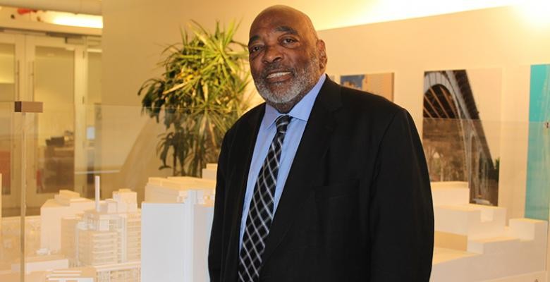 Wilmouth Elmes dressed in dark suit with light blue shirt and dark tie in the lobby of Manhattanville location with model of architectural layout behind him