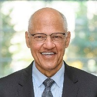 A bald, bespectacled man smiling, wearing a grey suit, blue buttoned-down shirt and gray-blue tie.