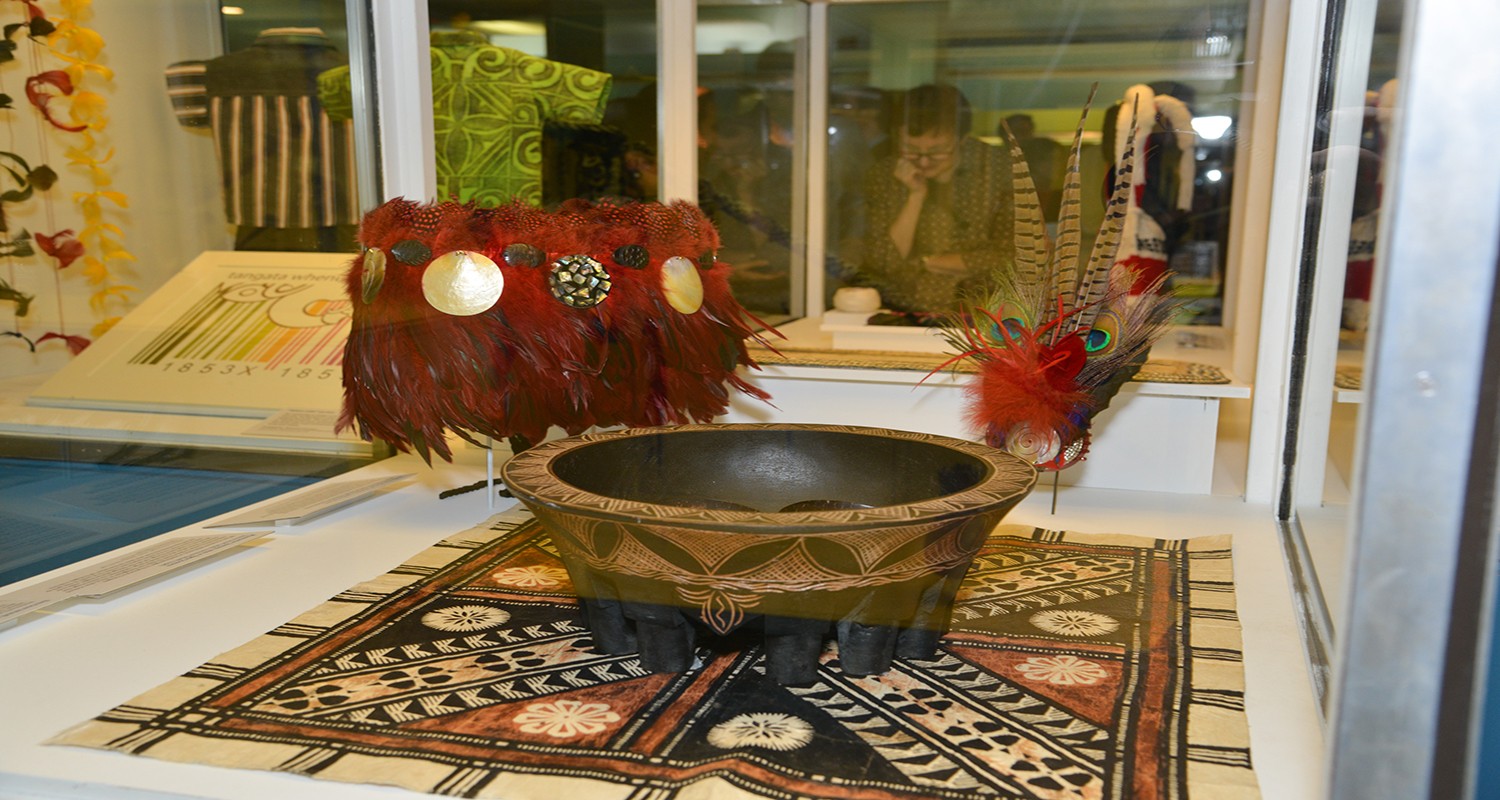 a feathered belt, bowl and headdress that are representative of Tongan culture. The bowl is sitting on a decorated Fijian masi, a barkcloth that is used in furnishings and clothing. 