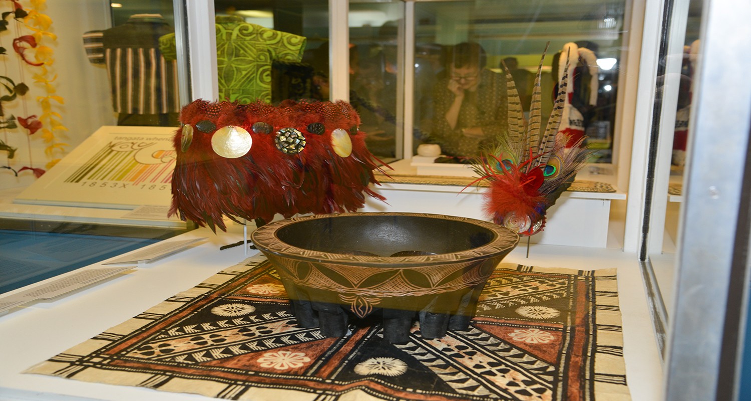 a feathered belt, bowl and headdress that are representative of Tongan culture. The bowl is sitting on a decorated Fijian masi, a barkcloth that is used in furnishings and clothing.
