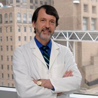 headshot of man with brown hair in white medical coat crosses arms with building in background