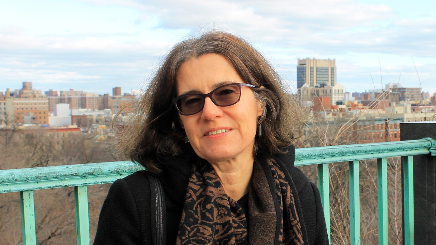 A woman wearing glasses, a scarf and a dark coat stands in front of a green railing.