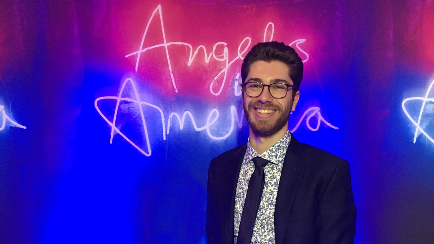 David Treatman: a bespectacled man with dark hair, in jacket, tie and floral shirt, standing in front of a pink and blue backdrop with the words "Angels in America" in cursive across the neon background.