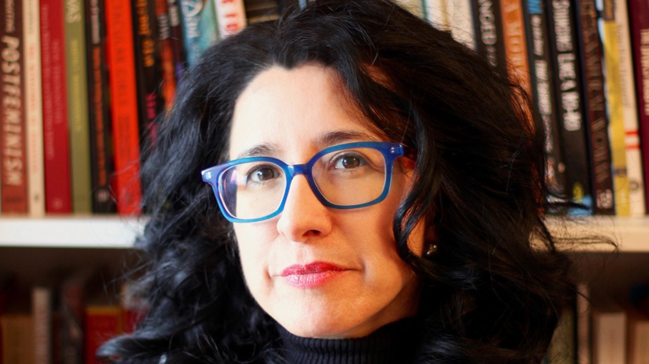 A woman wears glasses and has dark wavy hair and stands in front of bookshelves.