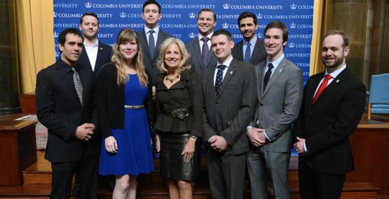 Dr Jill Biden wears a black leather skirt with a black blouse and necklace. She is located in the middle of several miliary veterans now students at Columbia University.  The men are dressed in suits and ties. The young lady is dressed in a royal blue dress and a black blazer while stands next to Dr. Biden.