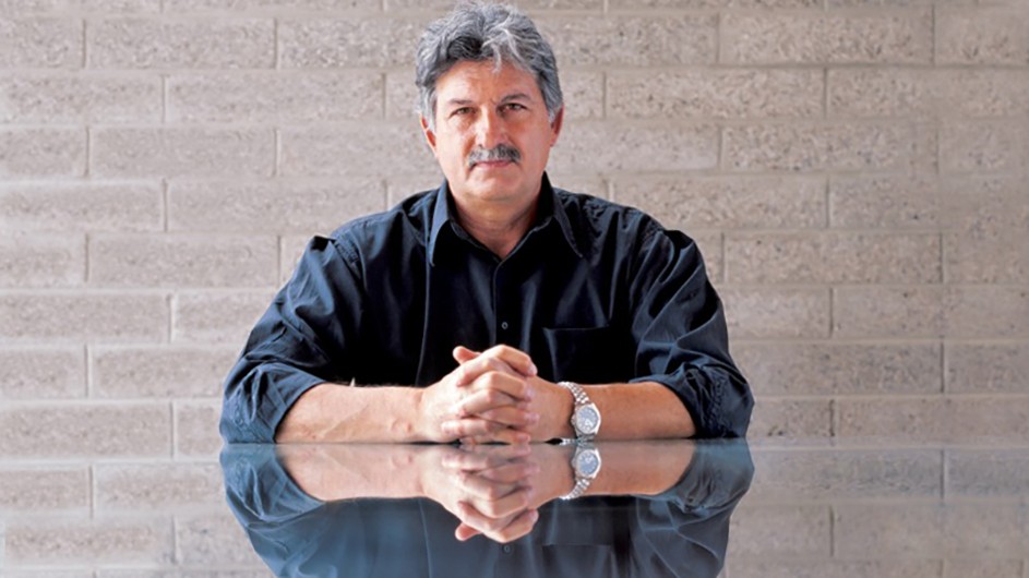 A man with a mustache wearing a dark shirt and a big watch sits at a table with folded hands in front of a brick wall.