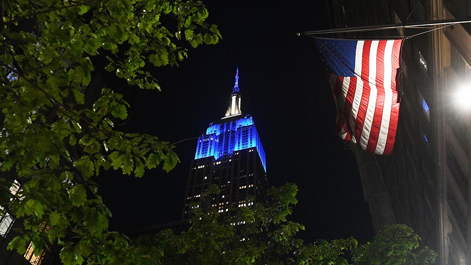 The Empire State Building in blue next to an American flag. 