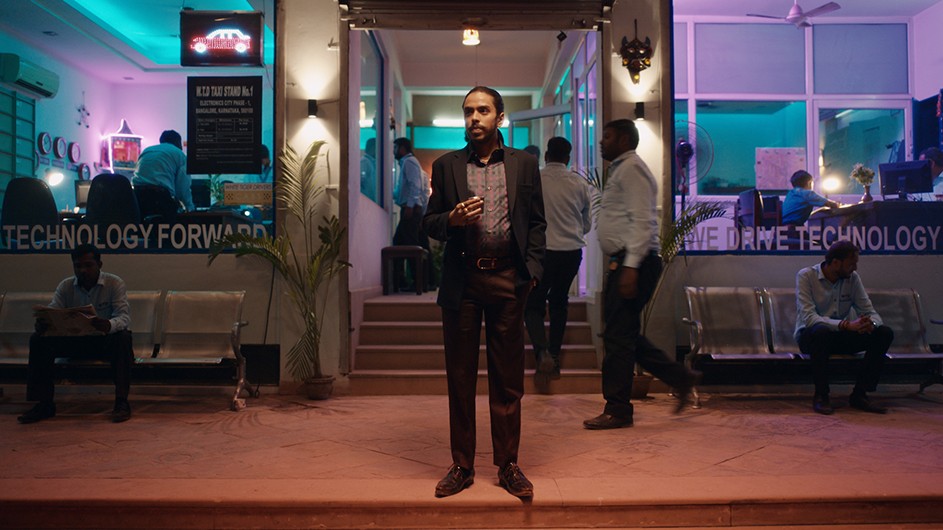 Adarsh Gourav plays Balram in "The White Tiger." Image courtesy of Neflix. A man stands on a sidewalk in front of passersby and a well-lit, glass-fronted store.