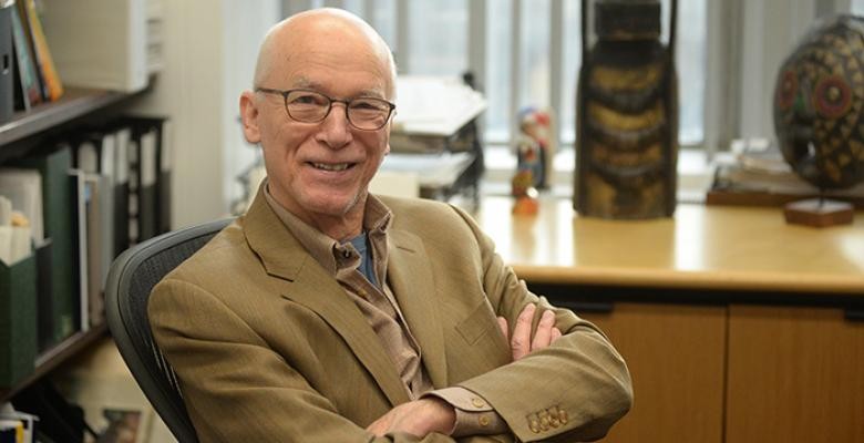 Prof Heal seated in front of a wooden desk wearing a tan colored blazer with a tan button down underneath and dark rimmed eyeglasses