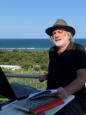A man with a white beard and wearing a hat sits at a table with books before a background with greenery and water.