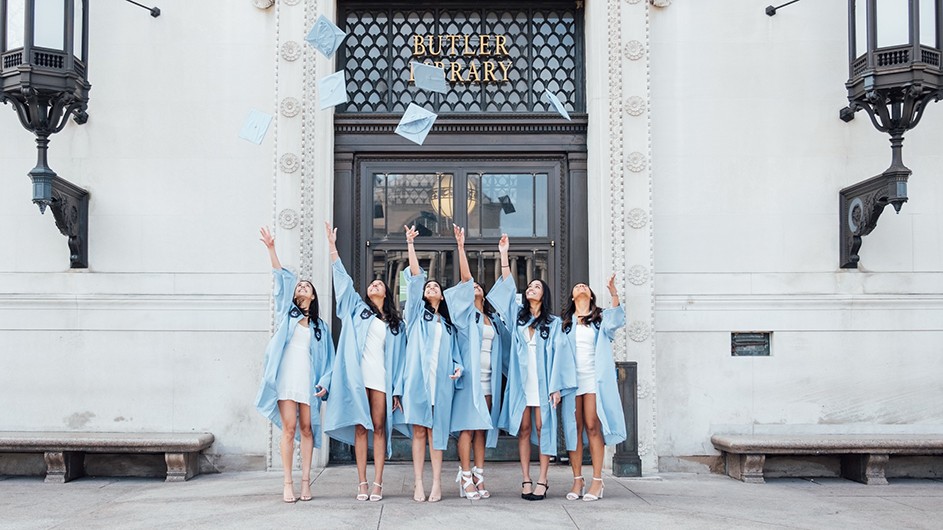 Six women throw their mortar boards in front of Butler Library in Columbia regalia. 