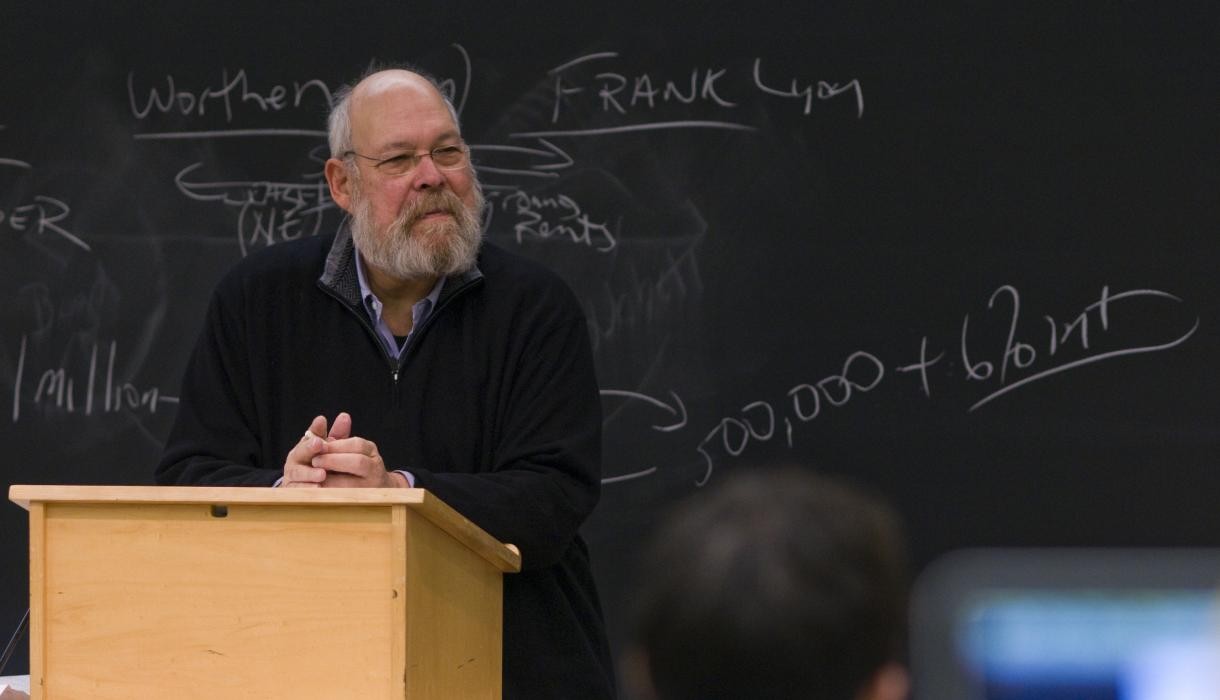 A man stands behind a lectern and in front of a blackboard.