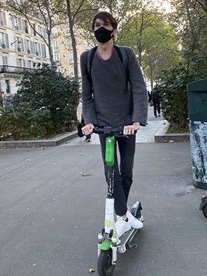 Ian William Christensen: A masked young man wearing dark clothes rides a scooter on a Paris street.