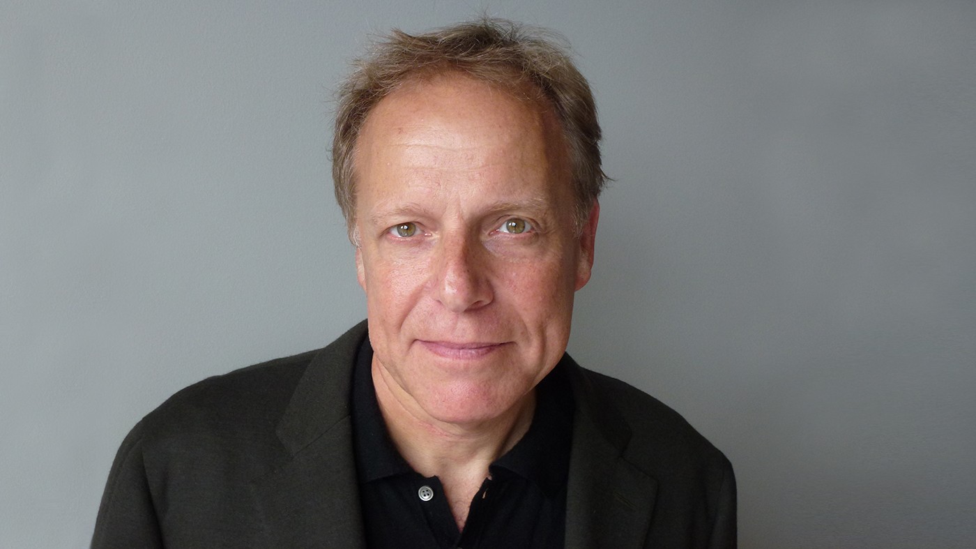 Headshot of James Shapiros: a man with blond hair, wearing a black shirt and black jacket poses against a gray background.