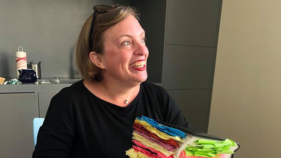 A smiling woman wears a dark shirt and holds a big book with colorful paper post-it notes stuck inside.